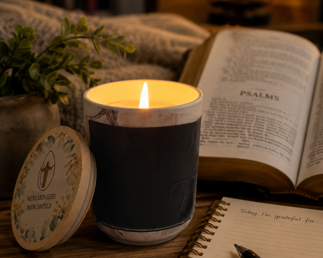 A lit candle in a white ceramic jar with a dark navy blue wrap stands on a wooden table beside its lid, which is printed with the scent name 'Mushroom Coffee'. In the background, there is an open book, a notebook with a pen, and a cozy indoor setting with warm lighting.
